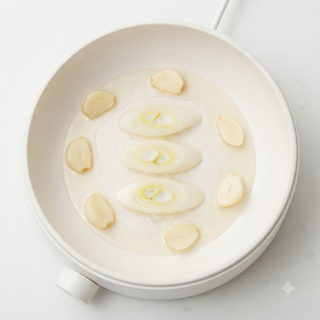 Green onion slices, garlic cloves, and potato chunks sizzling in oil inside a white cooking pot.
