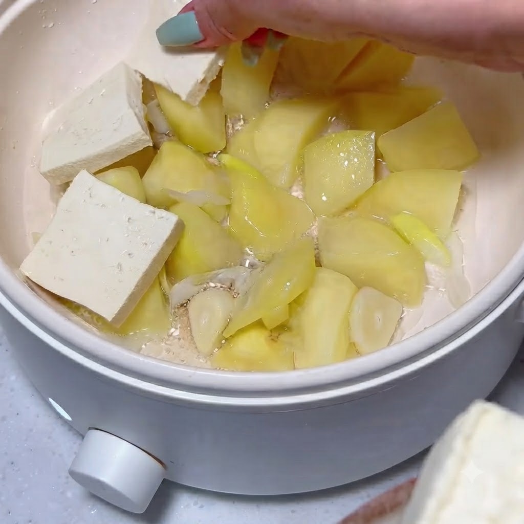 Tofu slices being neatly arranged over a layer of cooked potato chunks and garlic inside a white pot.