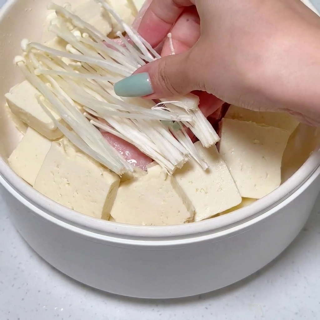 Enoki mushrooms being placed into the pot among arranged tofu and luncheon meat slices.
