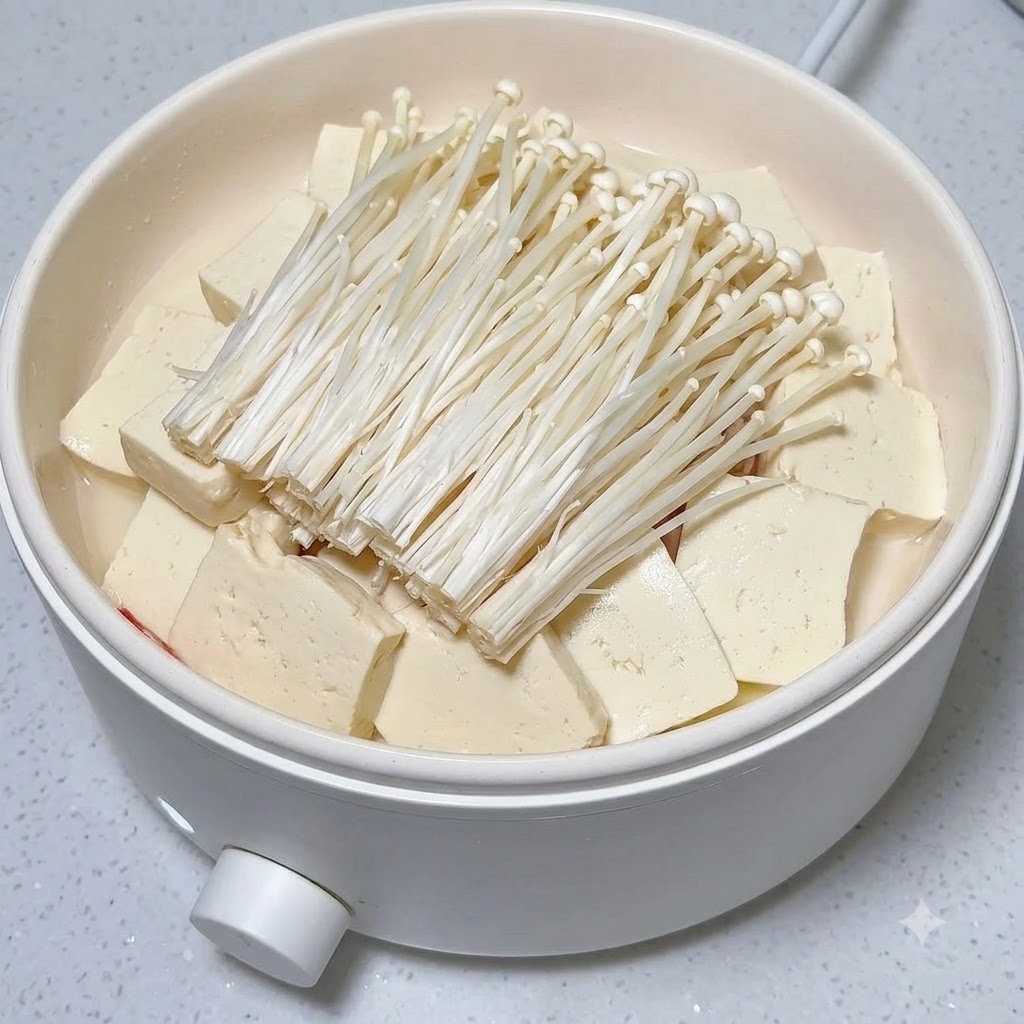 Fresh red chili slices neatly arranged on top of a heap of enoki mushrooms and tofu inside a white pot.
