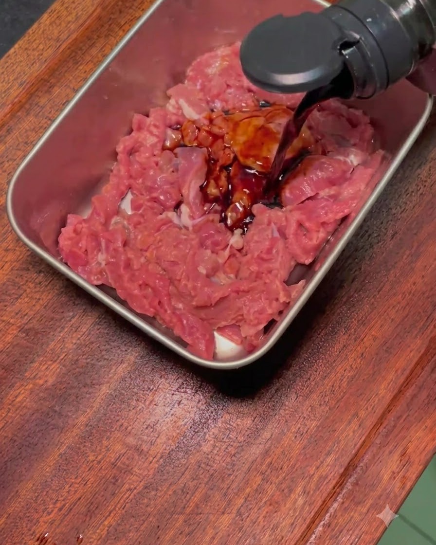 Dark marinade sauce being poured over raw, sliced beef in a rectangular metal tray.
