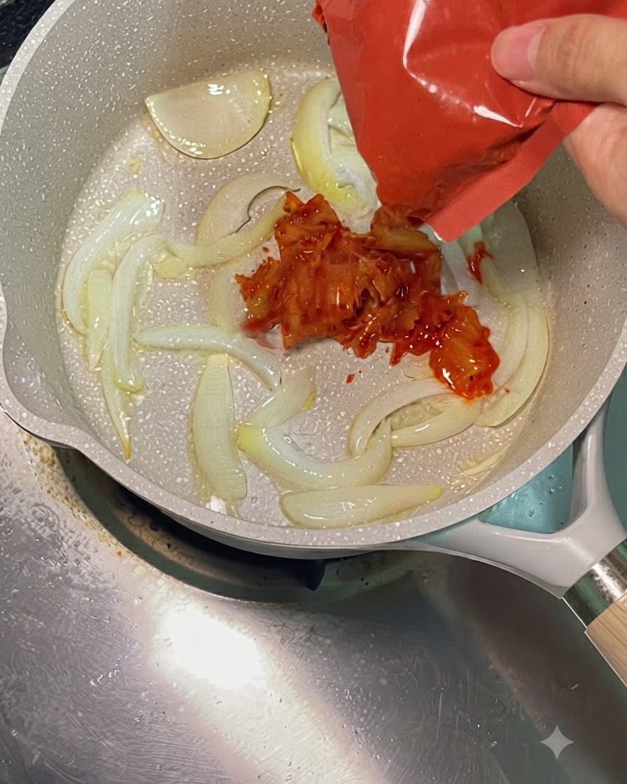 Bright red chopped kimchi being added from a red package into a hot pan with sautéed sliced onions.