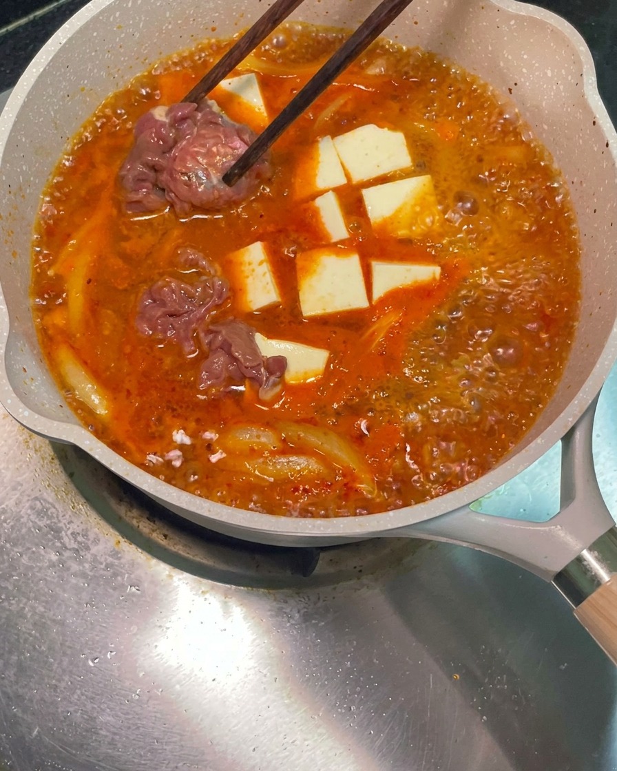 Chopsticks placing raw marinated beef slices into the simmering red soup pan.