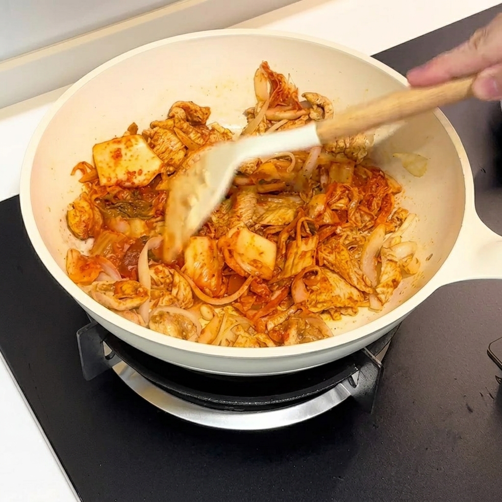 Kimchi and chicken being stir-fried together in a white pan using a spatula.