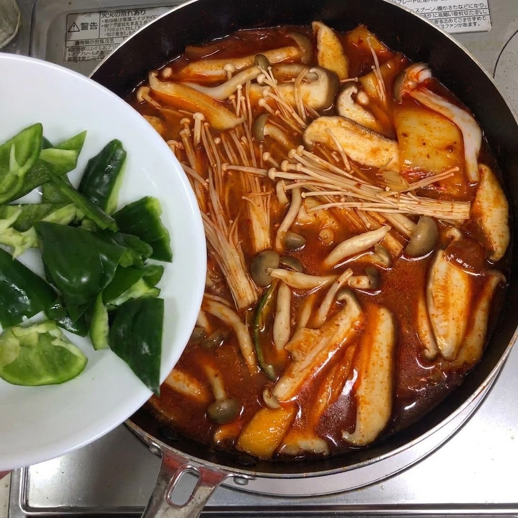 Slices of green peppers being added from a white plate into a pot of red kimchi soup.