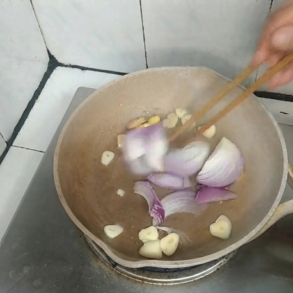 Sliced red onions, garlic, and ginger being stir-fried together in a pan using wooden chopsticks.