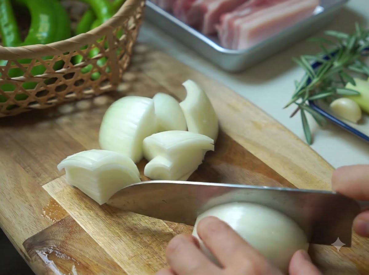 An onion being chopped into chunks next to a basket of green peppers on a wooden board.