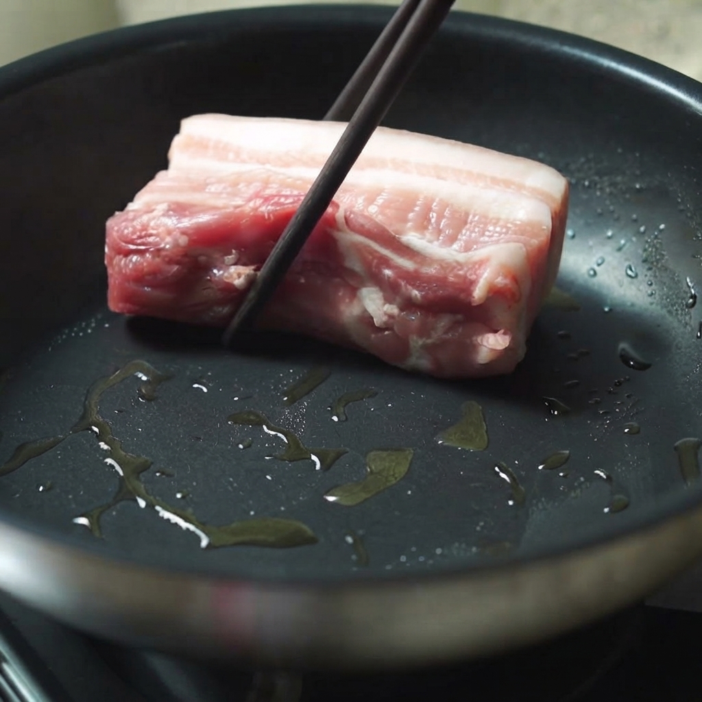 A thick piece of pork belly being placed into a hot oiled frying pan using chopsticks.