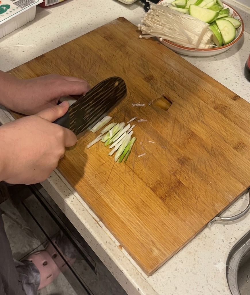 Hands using a kitchen knife to slice green onion stalks on a wooden cutting board.