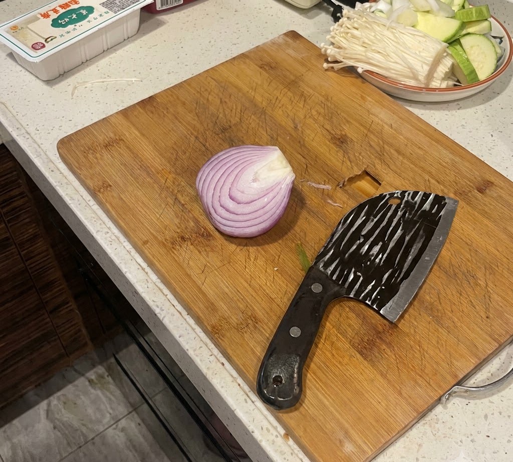 A halved red onion sitting on a wooden cutting board next to a large cleaver.