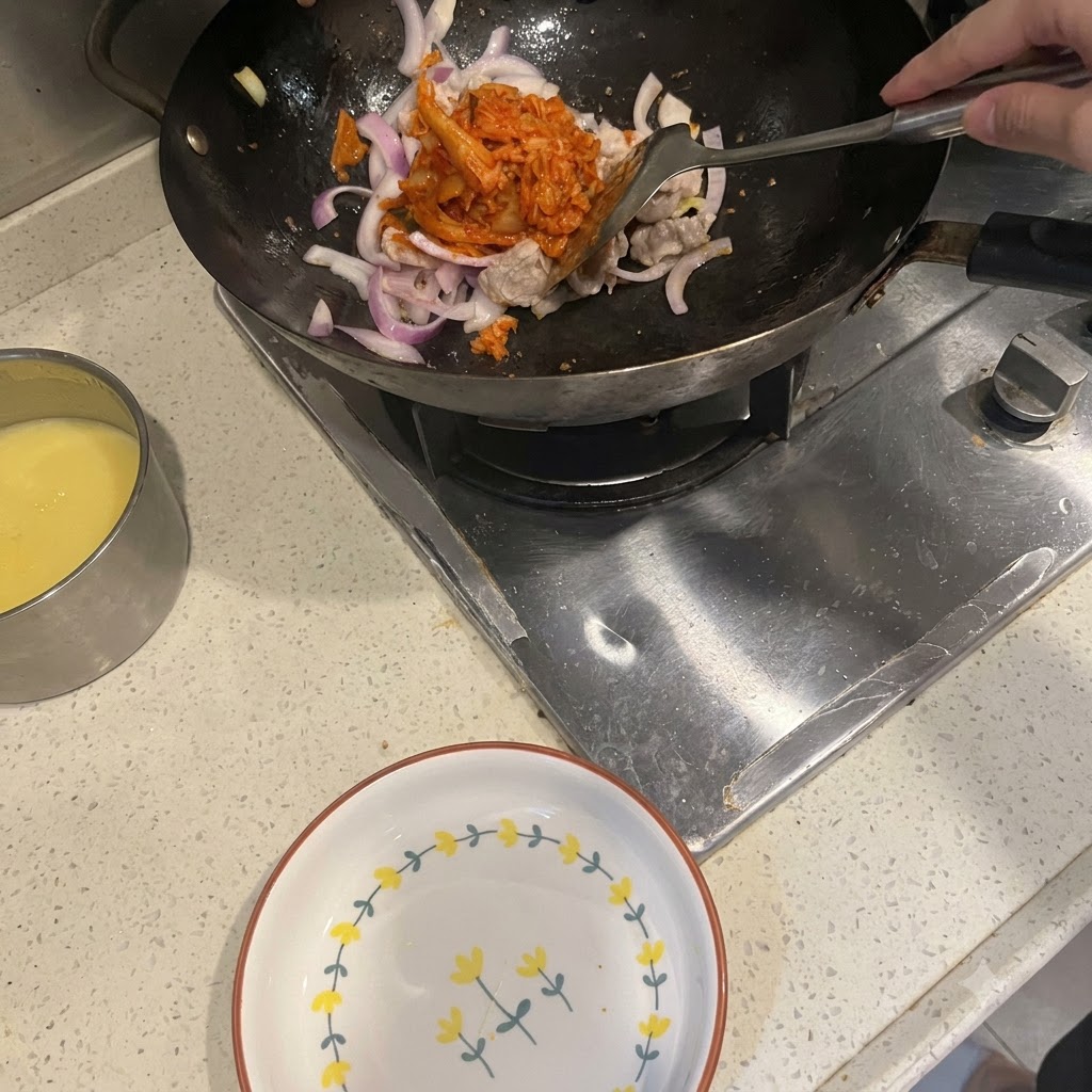 A spatula adding bright red kimchi into a wok containing stir-fried pork belly and red onions.