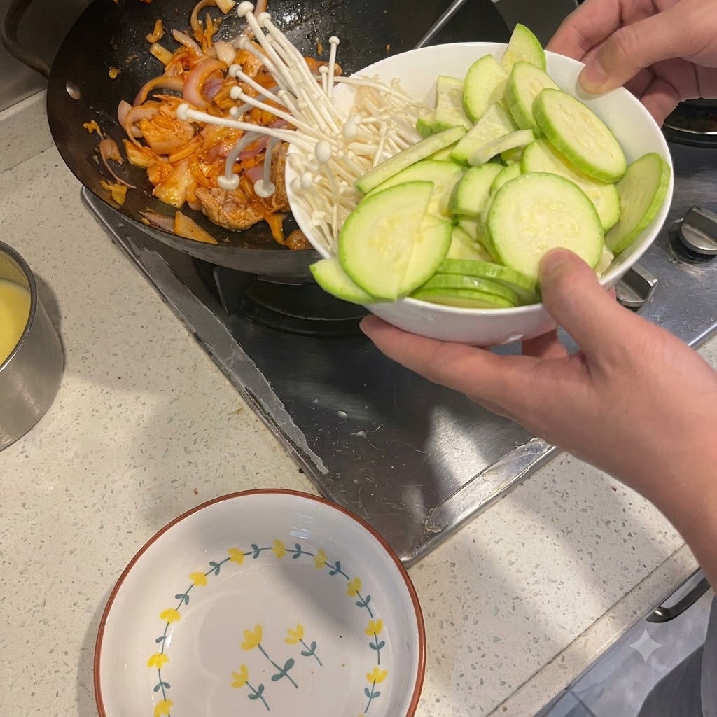 Sliced zucchini and enoki mushrooms being tipped from a bowl into a wok containing stir-fried pork and onions.