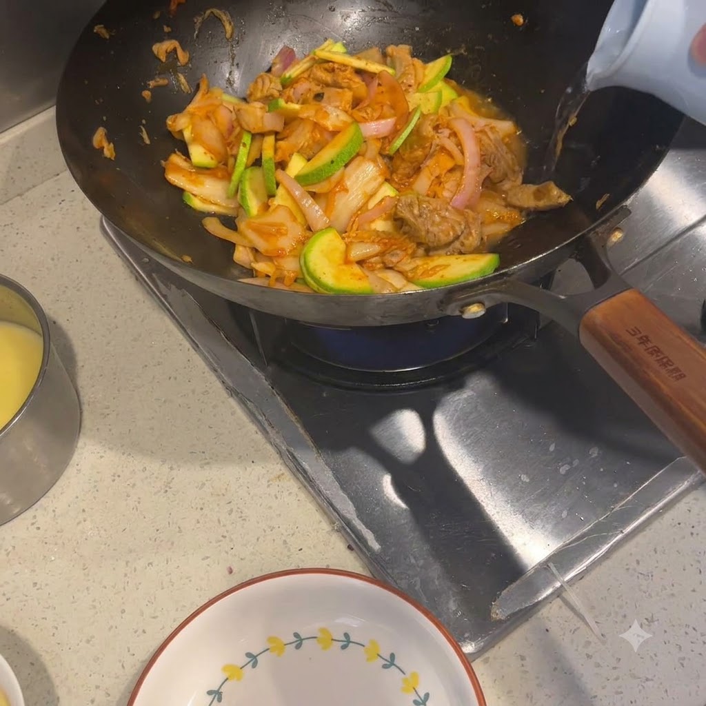 Hot water being poured from a light-colored kettle into a wok filled with stir-fried ingredients.