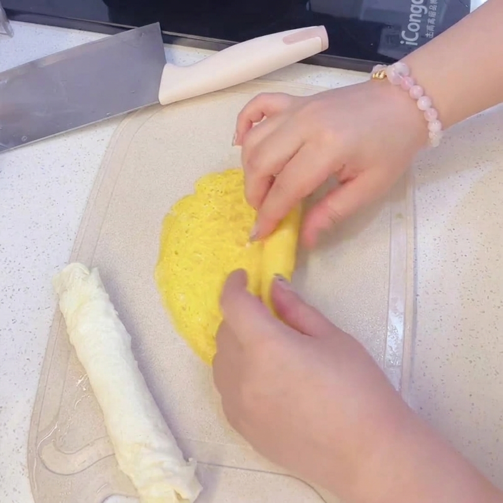 Hands rolling a thin yellow egg crepe on a cutting board, next to a pre-rolled white egg crepe.