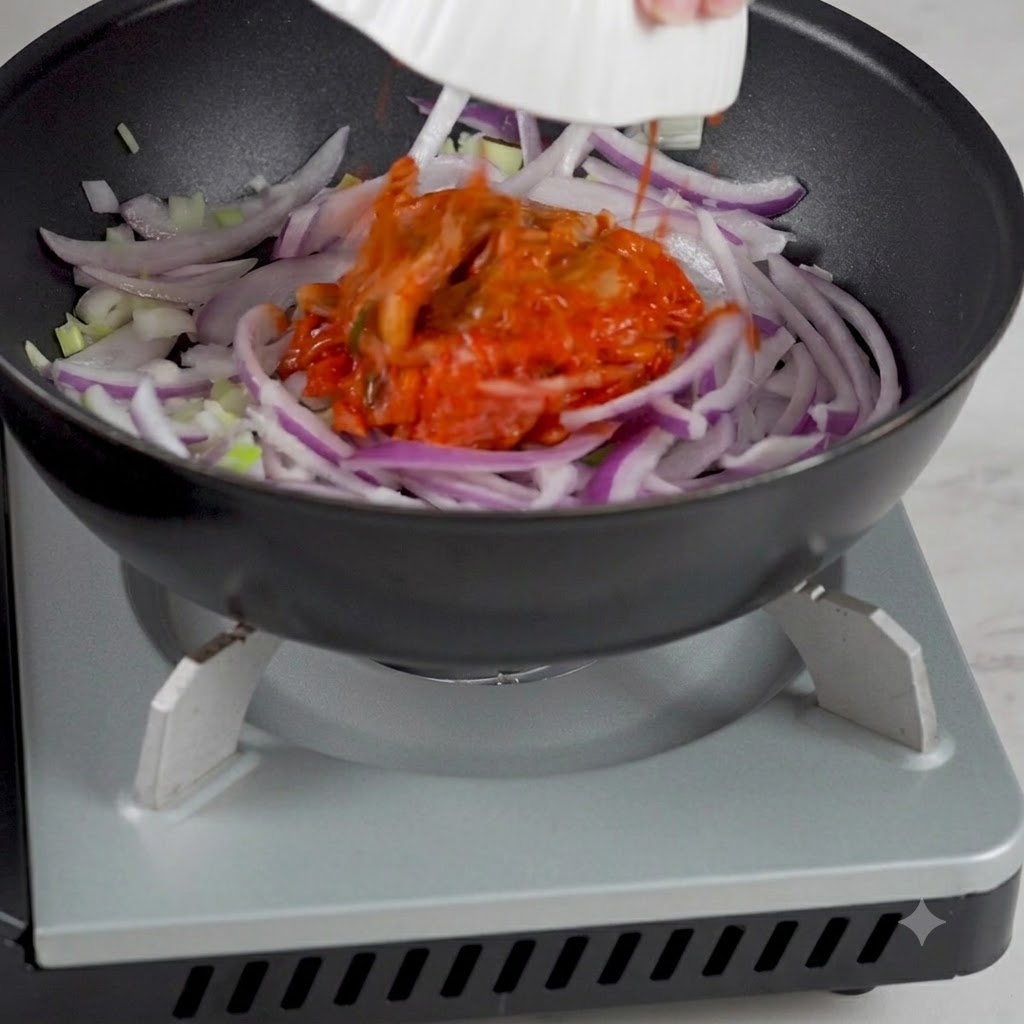 Korean kimchi being added from a bowl into the pan containing sautéed onions.