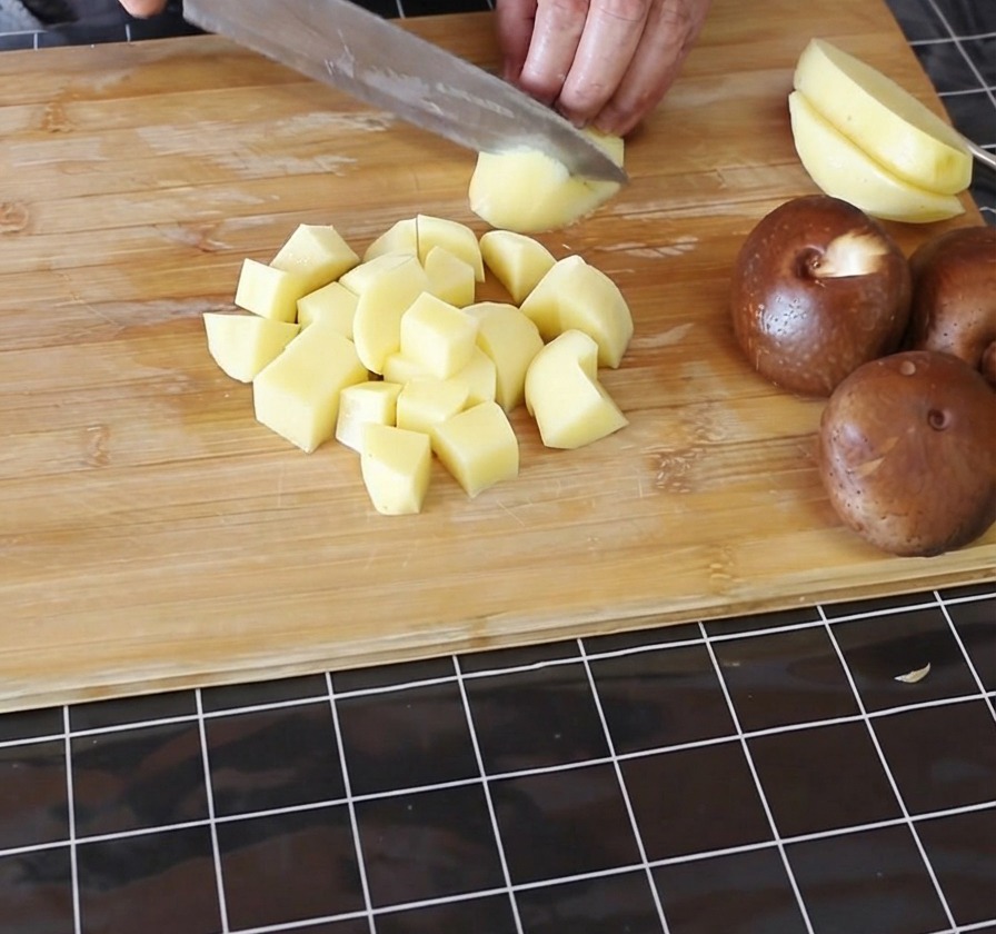Hands dicing peeled yellow potatoes into small cubes on a bamboo cutting board.