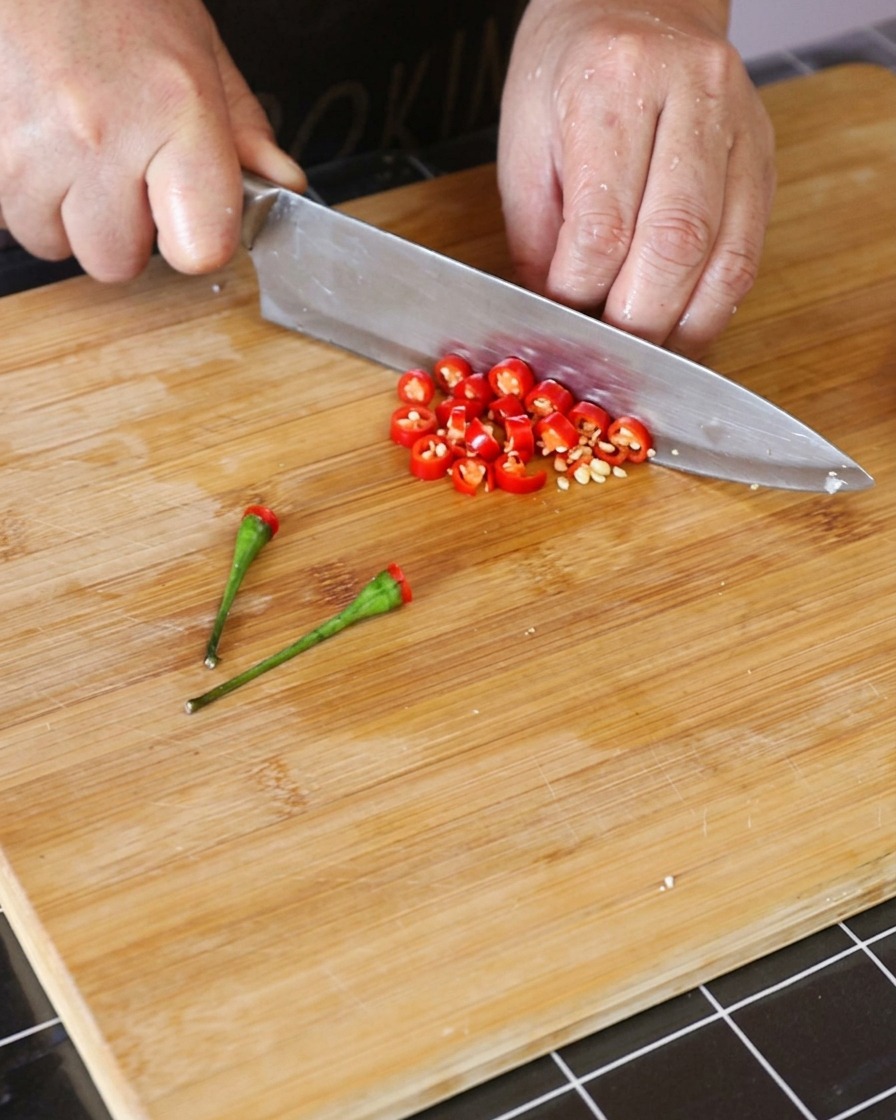 Fresh red Thai chili peppers being sliced into small rings on a wooden cutting board.