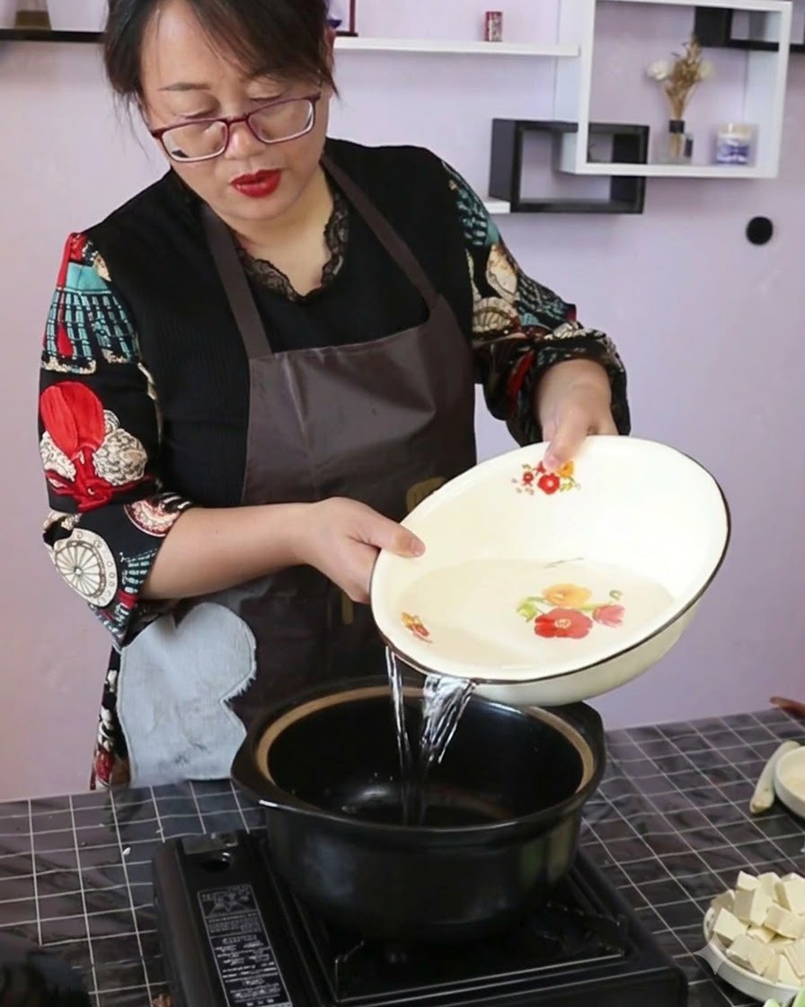 Clear water being poured from a decorative bowl into a black ceramic cooking pot on a portable stove.
