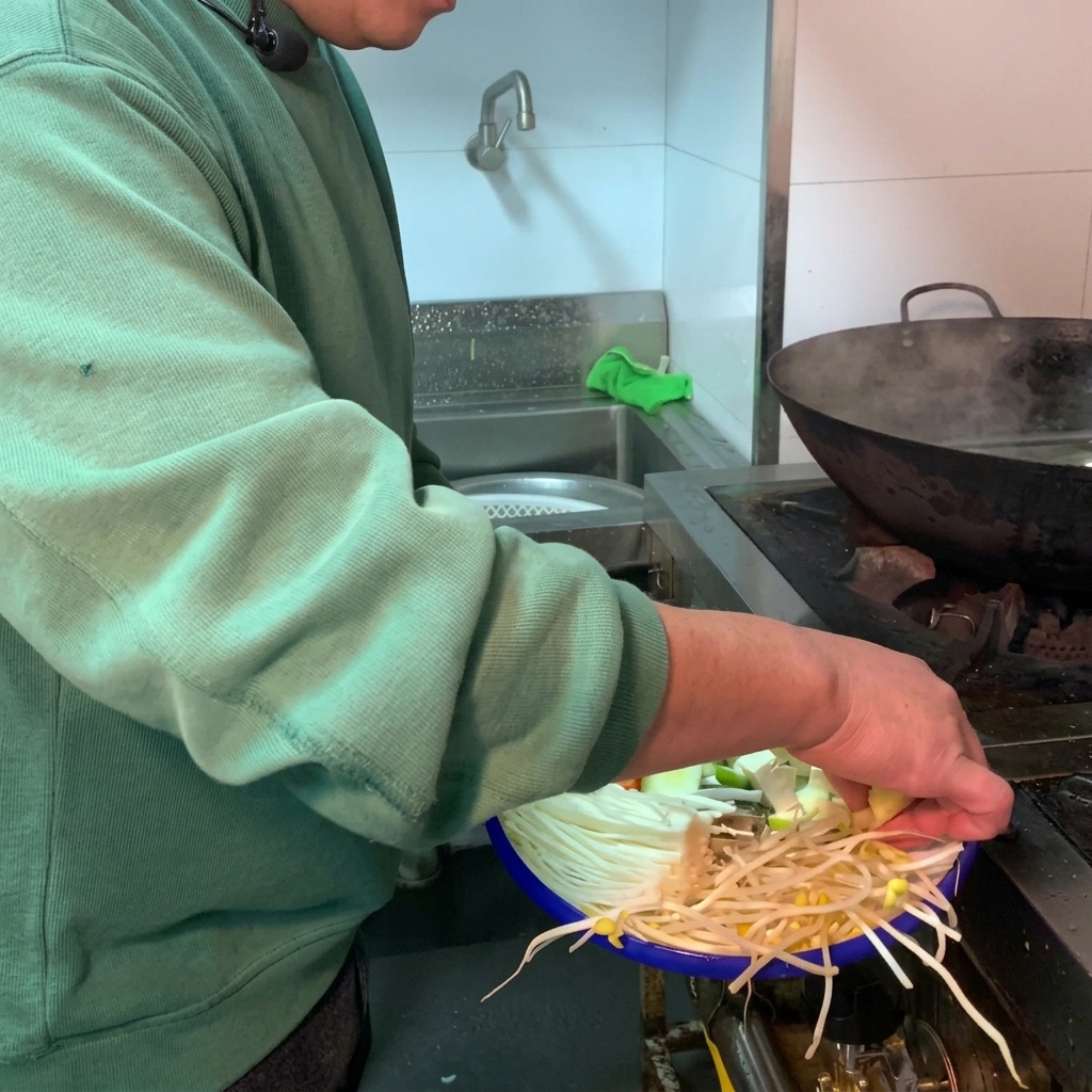 A variety of fresh vegetables and white tofu being added into a dark earthenware pot filled with water.