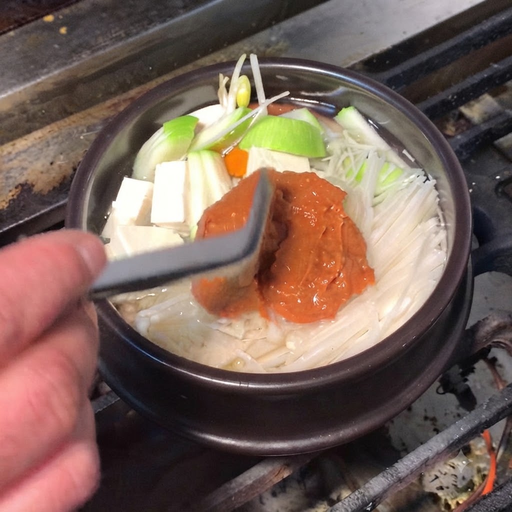 A silicone spatula adding rich brown soybean paste and red chili paste into the cooking pot.