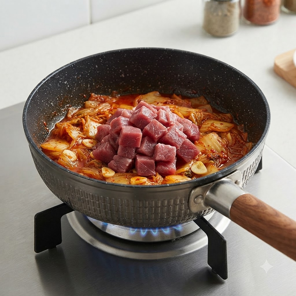 Raw beef cubes placed on top of stir-fried kimchi in a pot.