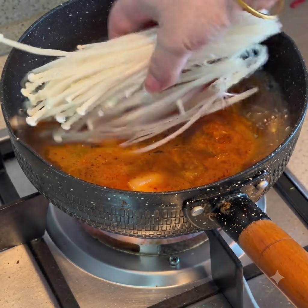Enoki mushrooms being added into a pot of boiling kimchi and beef stew.