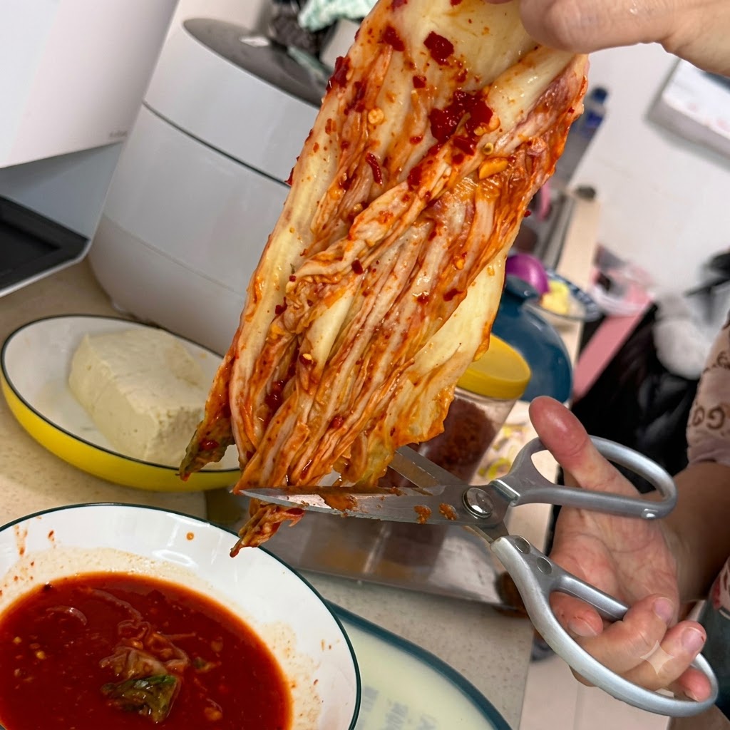 A pair of silver kitchen shears cutting a piece of red kimchi over a white bowl filled with spicy red sauce.
