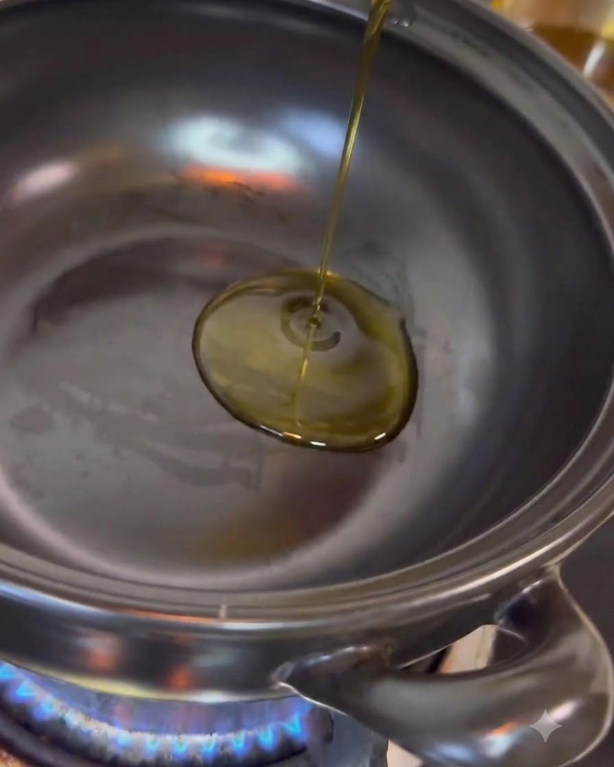 Clear cooking oil being poured into the center of a stainless steel cooking pot sitting on an active blue flame.