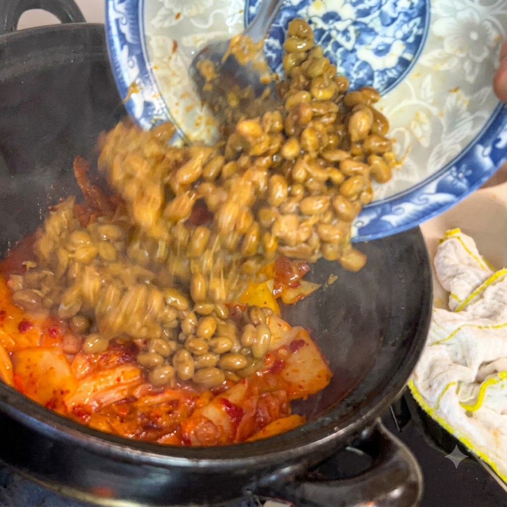 Fermented soybeans sliding from a blue and white decorative plate into a dark cooking pot filled with sizzling kimchi.