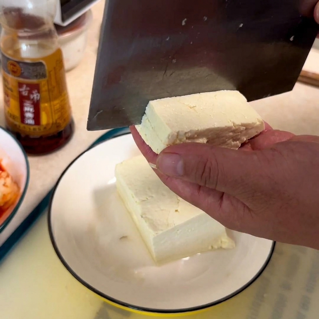 A hand holding a block of white tofu while a cleaver slices it into cubes on a white plate.