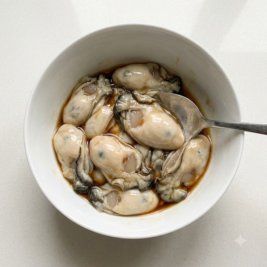 Cleaned oysters in a white bowl being seasoned with fish sauce and white pepper powder using a spoon.