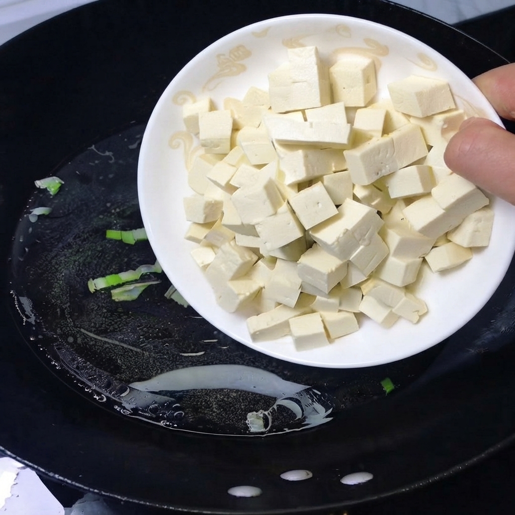 A plate of cubed white tofu being poured into a black wok with boiling water.