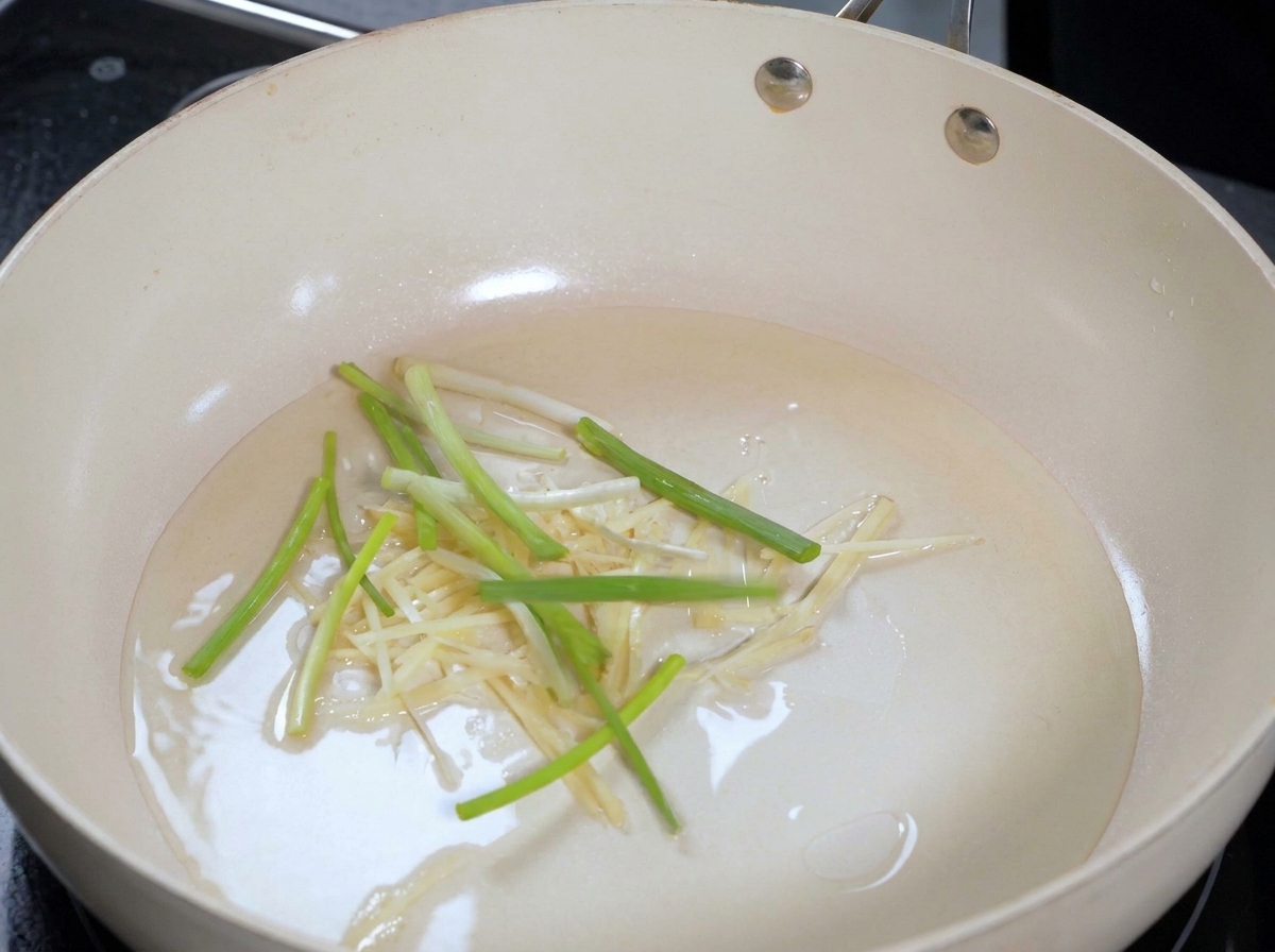 Sliced scallions and ginger strips sizzling in light oil inside a modern cooking pan.