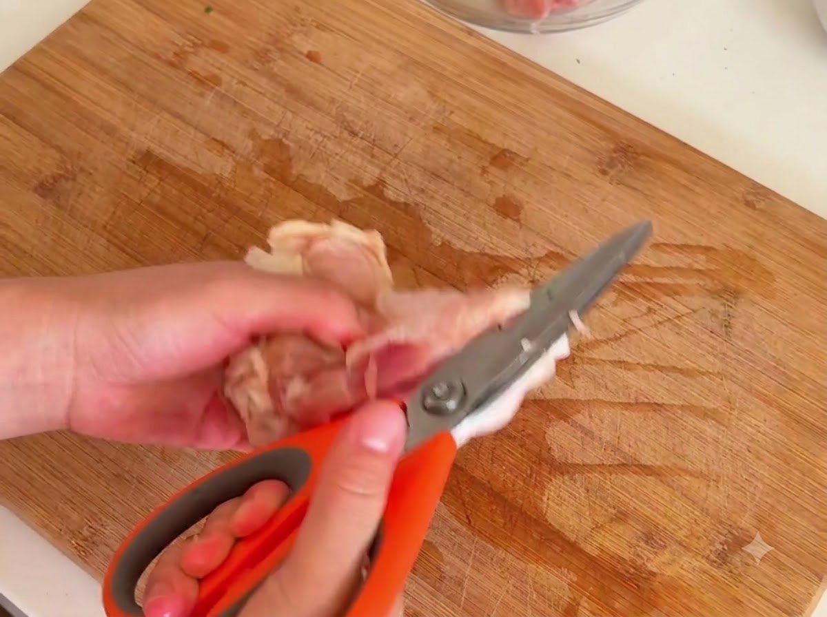 A pair of orange kitchen scissors cutting away the meat from a raw chicken leg bone on a wooden board.