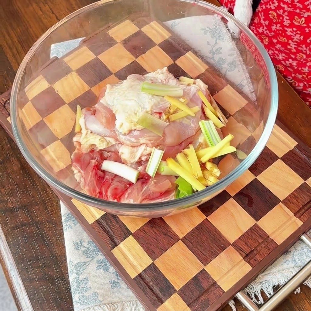 Raw chicken pieces in a glass bowl topped with sliced red onions, scallions, and matchsticks of ginger.