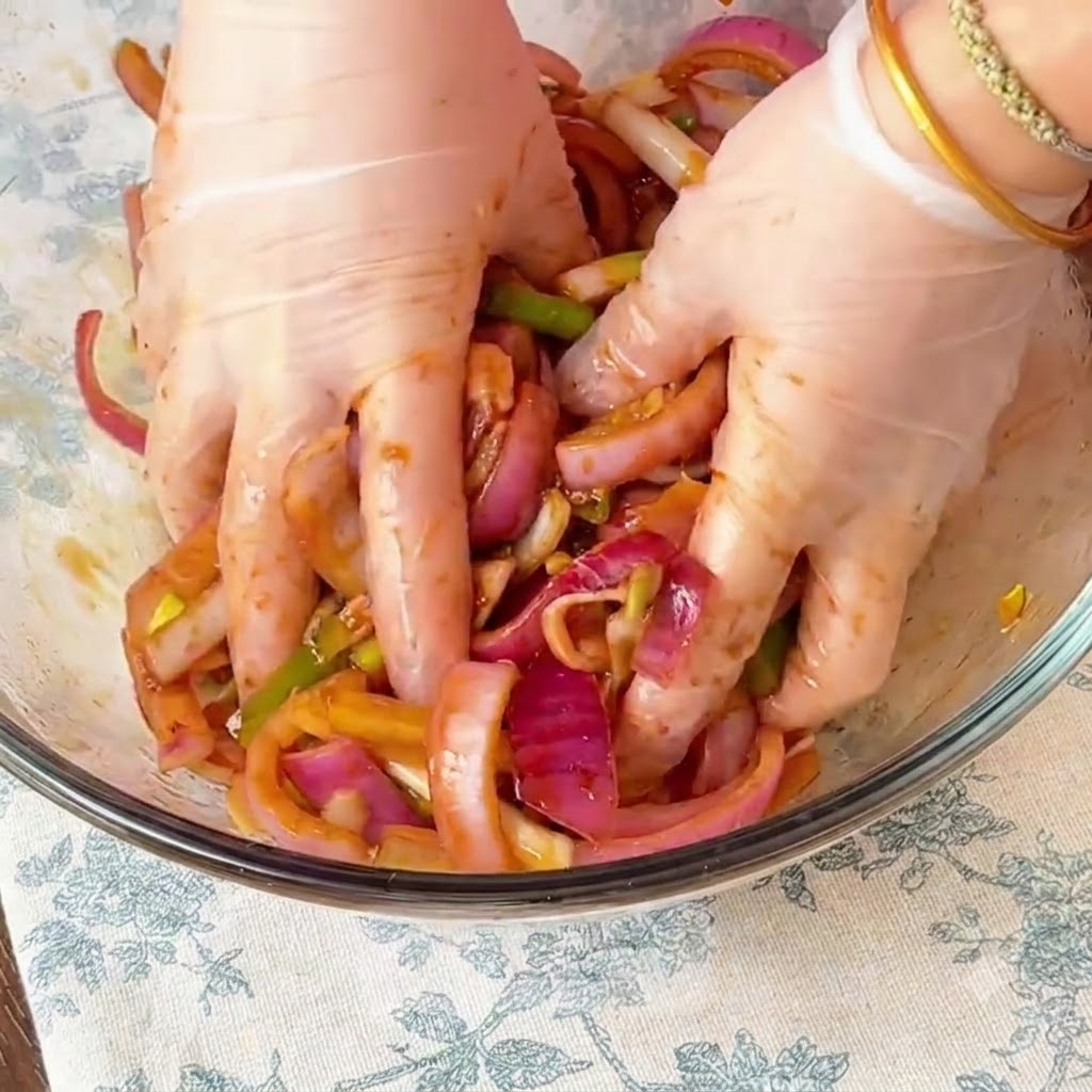 Gloved hands thoroughly massaging the marinade and sliced red onions into the chicken inside a glass bowl.