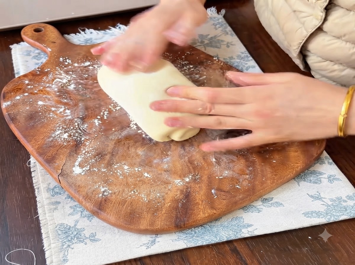 Hands rolling and shaping a portion of soft bread dough on a flour-dusted wooden cutting board.