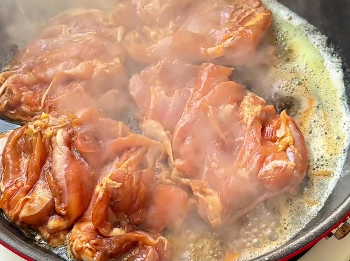 Marinated raw chicken thigh pieces sizzling in a hot frying pan with butter and steam rising.