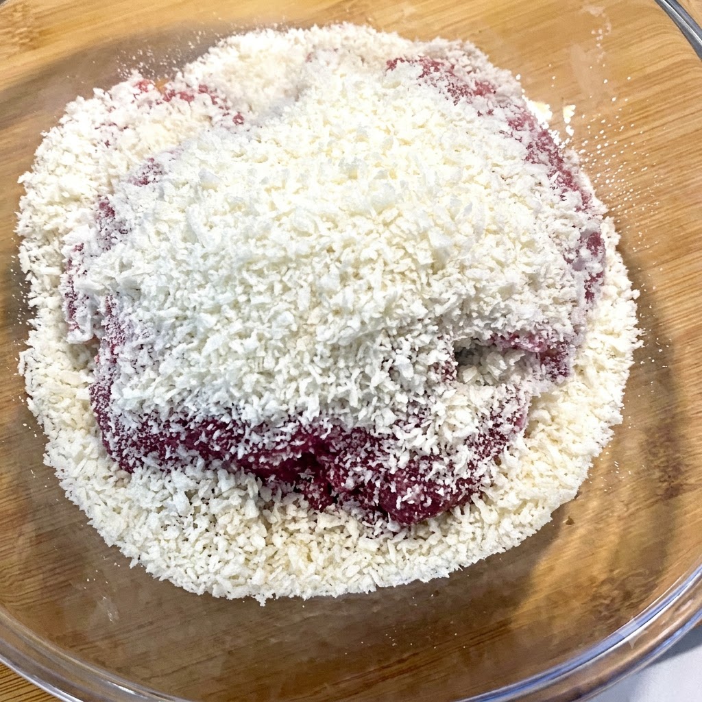 White breadcrumbs falling from a bowl directly onto raw minced beef in a glass container.