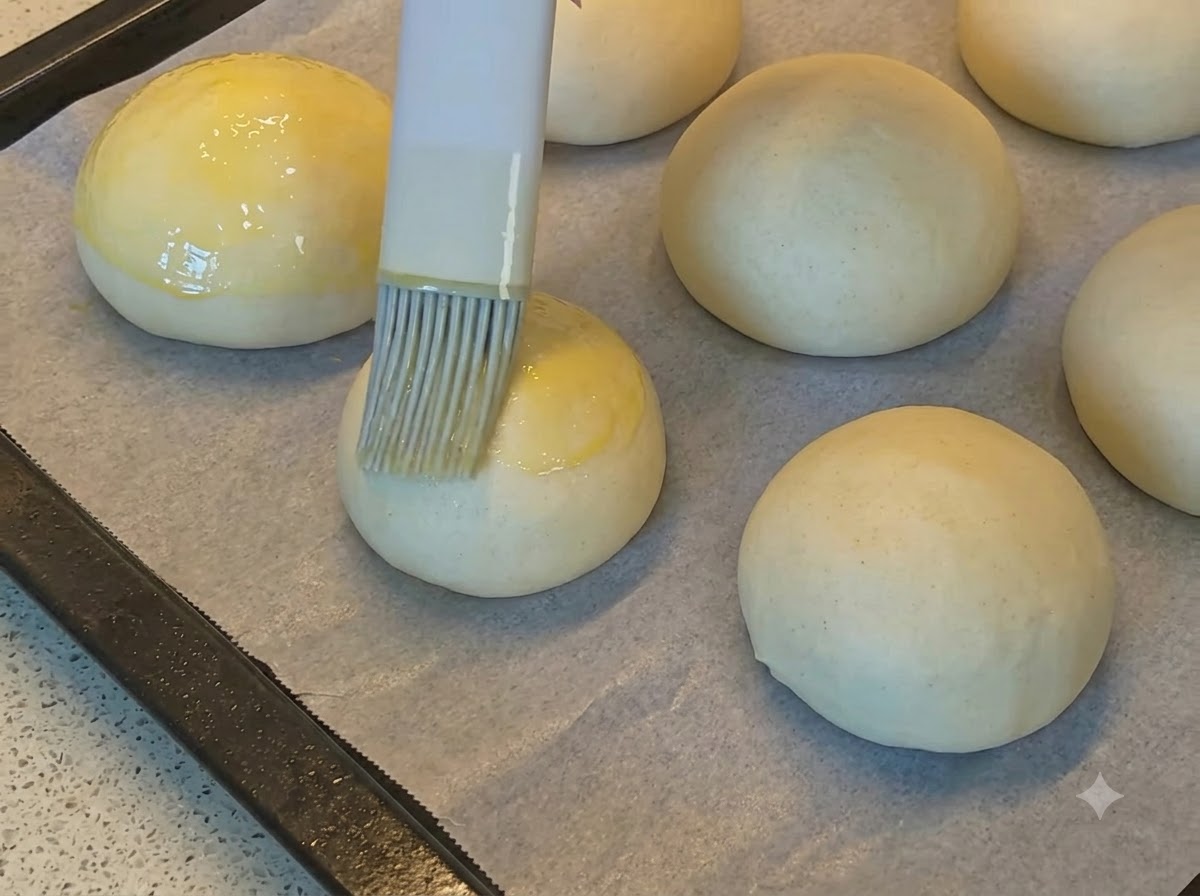 A pastry brush applying golden egg wash onto smooth round bread dough balls on parchment paper.