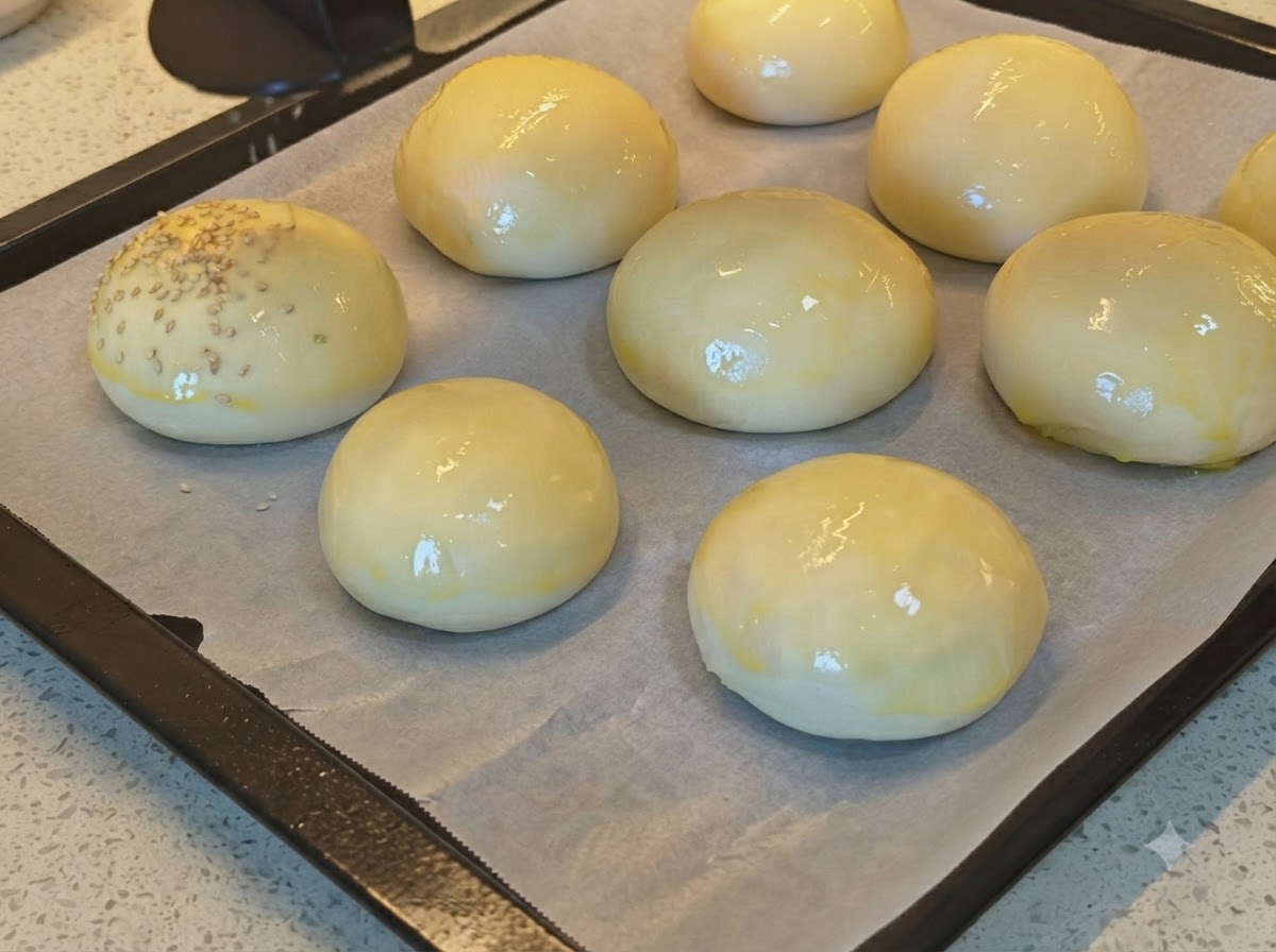 Sesame seeds being sprinkled onto a tray of shiny egg-washed bread buns.