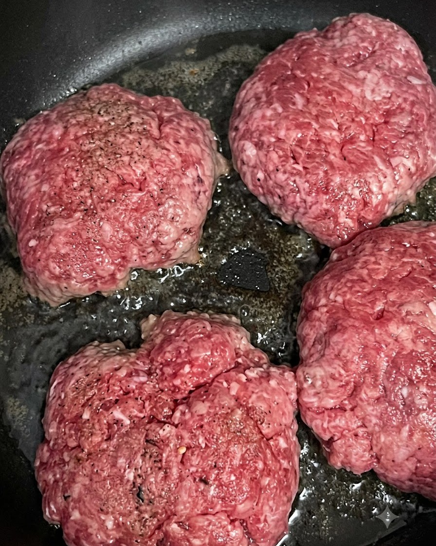 Four raw beef patties sizzling and browning in a hot frying pan.