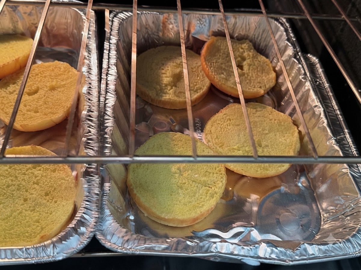 Burger bun halves resting on foil trays inside the oven heating coils.