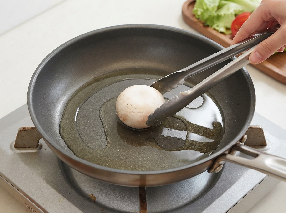 A single mushroom being placed into an oiled frying pan with tongs.