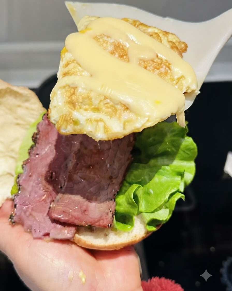 A spatula lifting a fried egg topped with melted string cheese over the burger assembly.