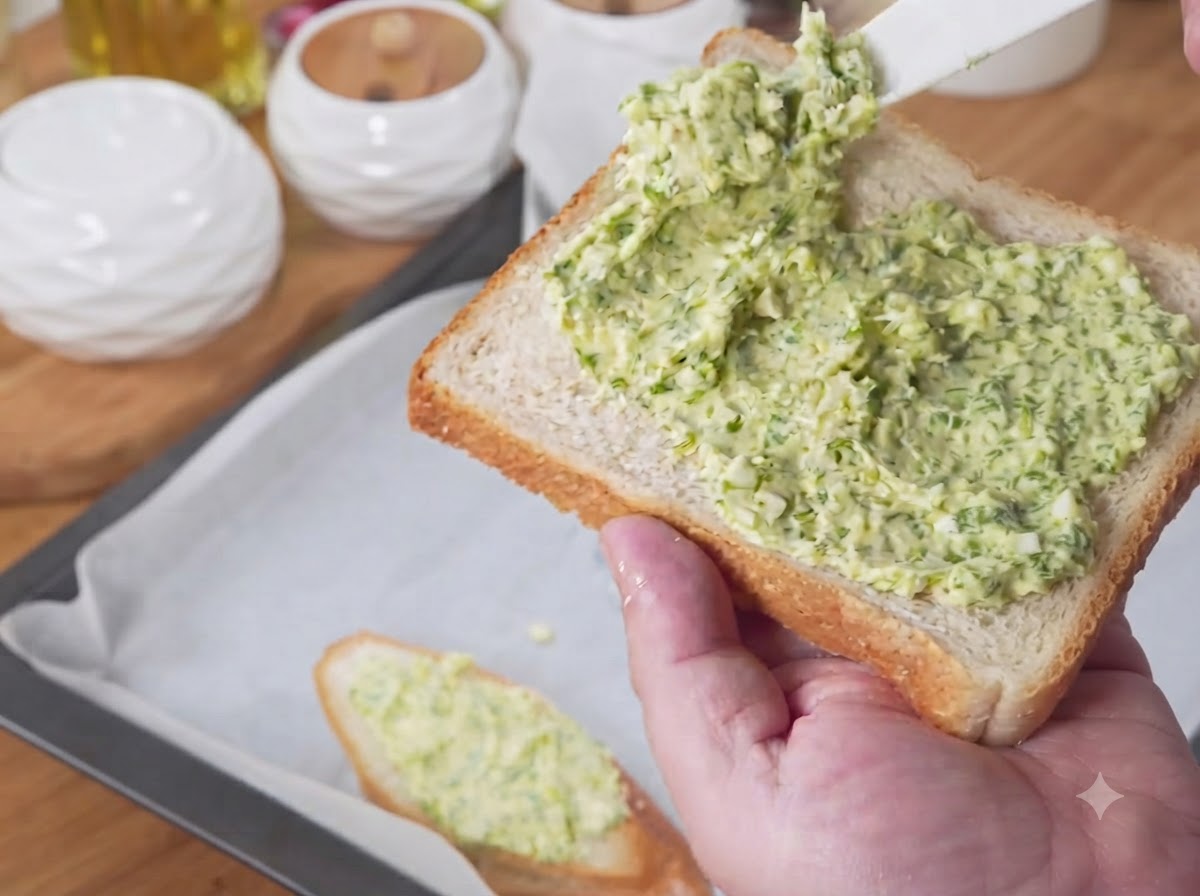 Slices of bread covered in green garlic butter mixture being arranged on a parchment-lined baking tray.