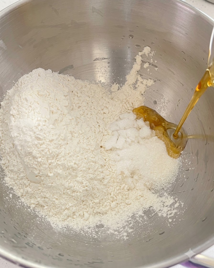 Dry flour, sugar, and liquid ingredients being poured together inside a metal stand mixer bowl.