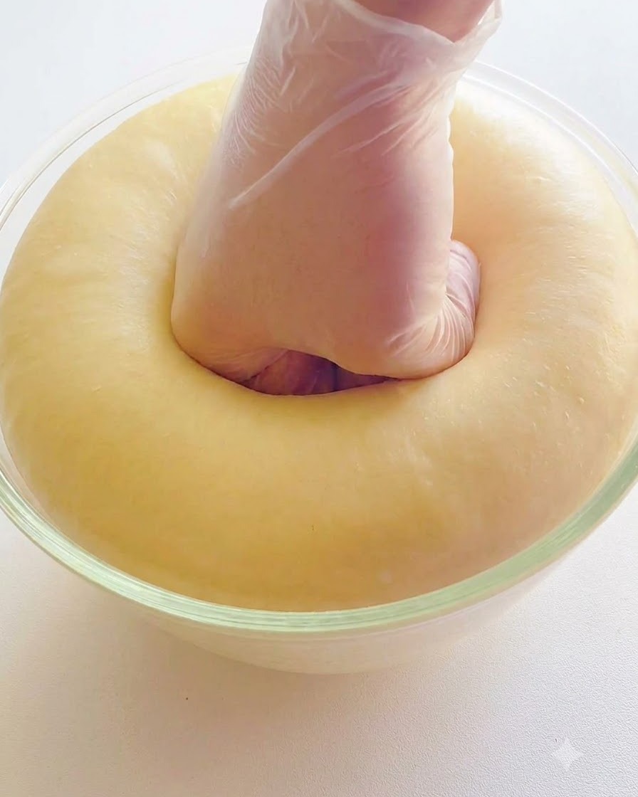 A hand in a clear glove punching down the center of a puffy, well-fermented dough in a glass bowl.