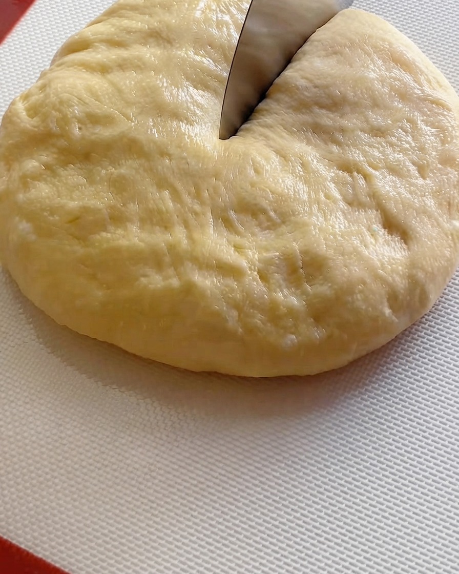 A dough cutter slicing a large fermented dough ball into nine portions on a white mat.