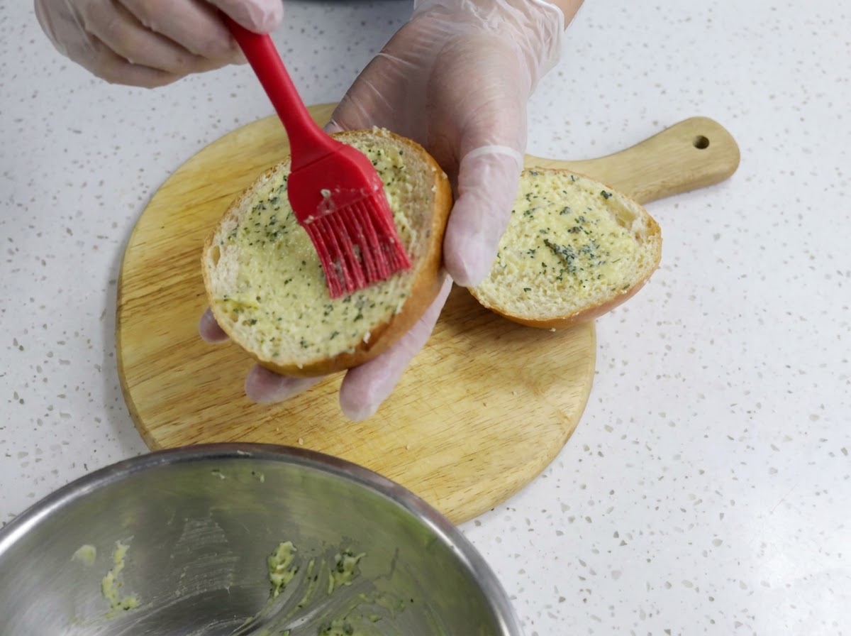 A gloved hand using a red silicone brush to spread garlic butter evenly over a cut burger bun half on a wooden board.